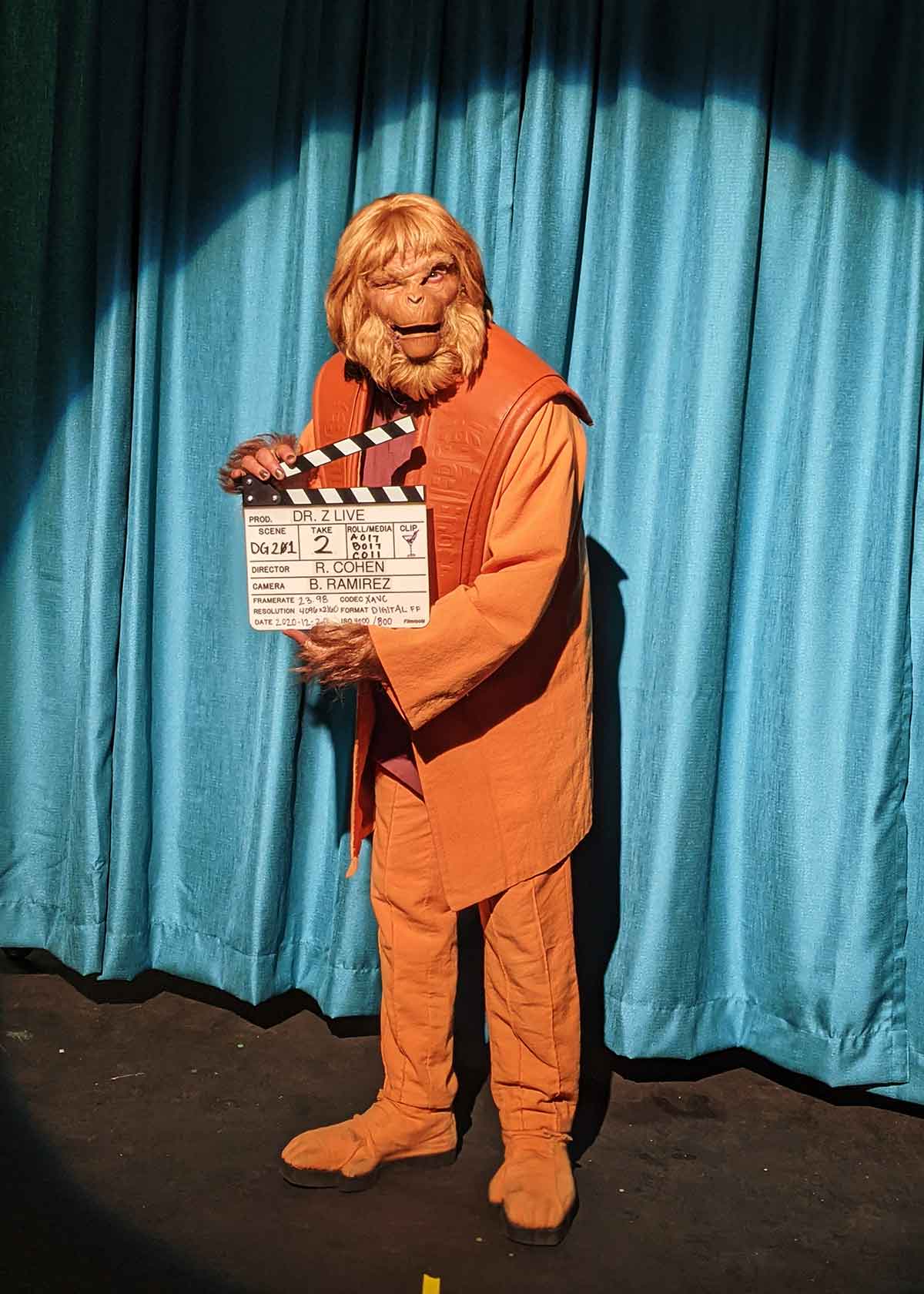 Dr Z in an orange costume holding a clapperboard against a blue curtain